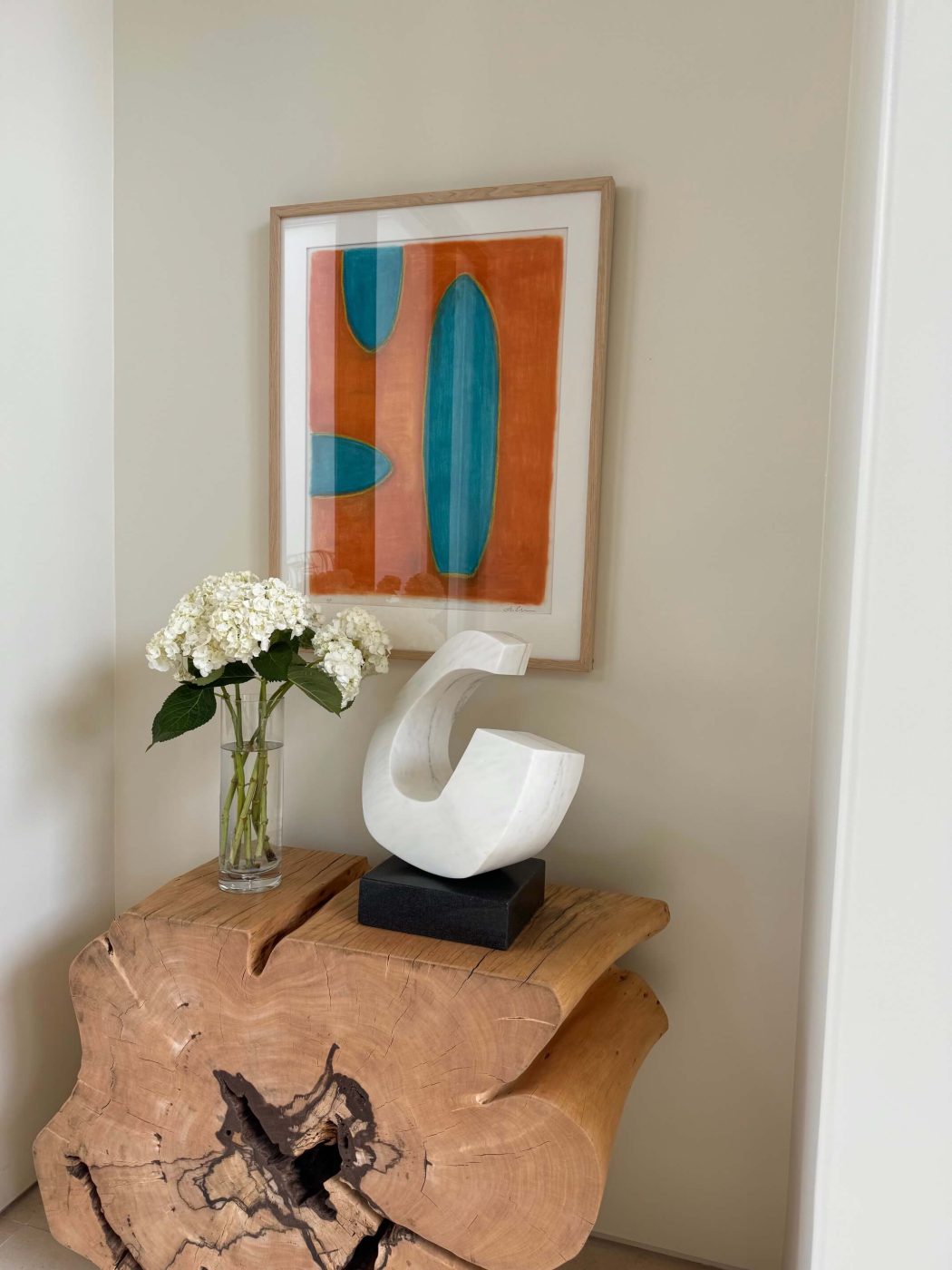 Orange and blue Lori Brunner painting above a wood-slab console with a white sculpture and flowers