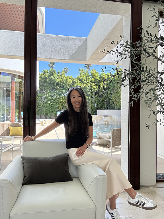 Lori Brunner seated on a white chair in her La Jolla living room, with the pool and garden behind her