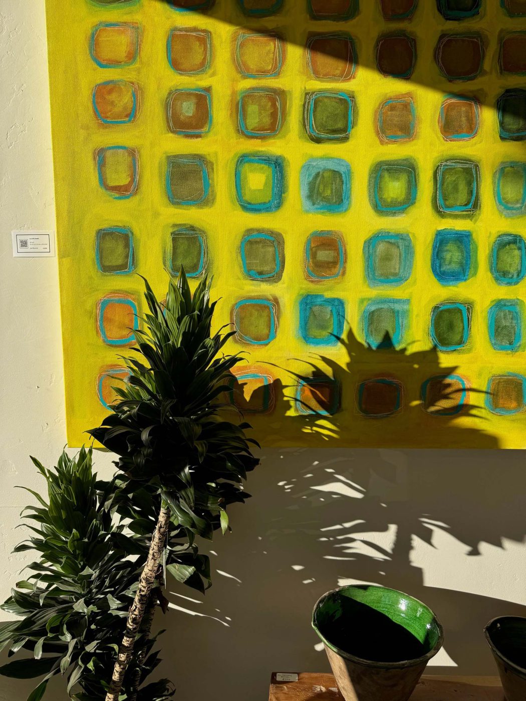 Large yellow and turquoise grid painting by Lori Brunner on a gallery wall, with a plant and green bowls in the sunlight